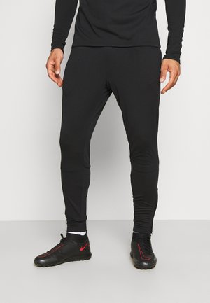 Mann trägt schwarze Sporthose, schwarzes Langarmshirt und schwarze Sneakers mit roten Nike-Logos, steht auf einem hellgrauen Boden.