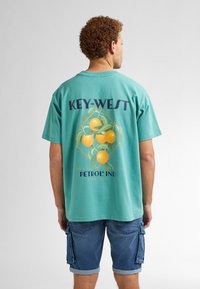 Turkoosi puuvillainen t-paita, jossa on graafinen kuvasto appelsiineista ja vihreistä lehdistä. Paidassa on tekstit "KEY-WEST" ja "PETROL IND" violetilla ja sinisellä.