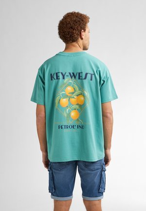 Camiseta de algodón turquesa con un diseño gráfico de naranjas y hojas verdes, con el texto "KEY-WEST" y "PETROL IND" en púrpura y azul.
