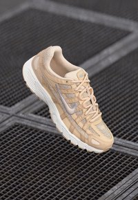Baskets beiges avec une tige en mesh et en daim, dotées d'une semelle blanche incurvée, d'accents gris et d'un système de laçage. Design léger et texturé.