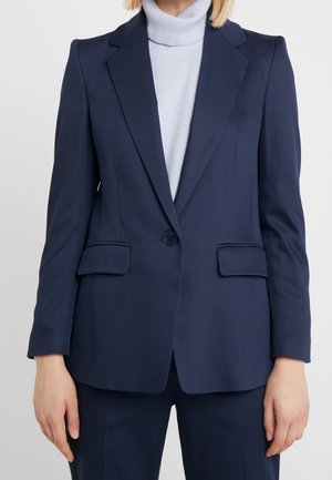 Frau trägt einen maßgeschneiderten dunkelblauen Blazer mit einem Knopf und Pattentaschen über einem hellgrauen Rollkragenpullover.