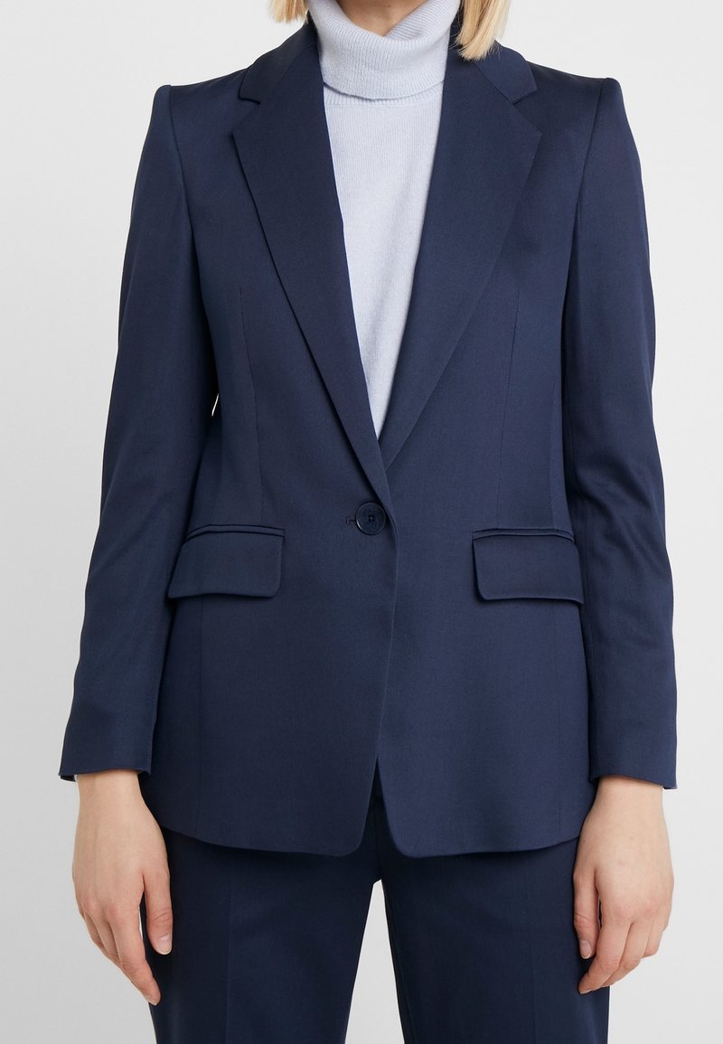 Femme portant un blazer bleu marine ajusté avec un bouton unique et des poches à rabat sur un pull à col roulé gris clair.