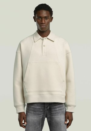 Lichtbeige sweatshirt met een kraagontwerp, voorzien van een voorkeurszak, geribbelde manchetten en een gestructureerde afwerking. G gedragen over donkere jeans.