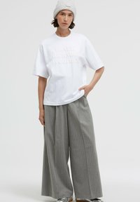White cotton oversized t-shirt with raised lettering, paired with wide gray pinstripe pants. Model wears a gray knit beanie and silver shoes.