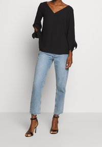 Femme portant un haut noir à col en V avec des manches en nœud, un jean bleu clair cropped avec des ourlets effilochés, et des sandales à talons hauts à bride de cheville noires.