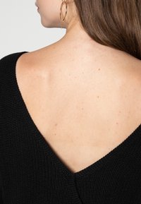 Maglione nero lavorato a maglia con ampio scollo a V, che mette in mostra un tessuto testurizzato. Caratterizzato da pelle liscia con lievi imperfezioni e un orecchino a cerchio dorato.