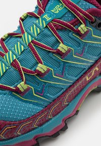 Sapatilha atlética turquesa com parte superior texturizada, acentos em marrom contrastante, atacadores verde néon e sola robusta. Detalhe padrão notável na entressola.