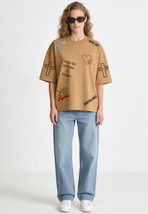 Junge Frau mit lockigem Haar, die schwarze Sonnenbrillen, ein beiges übergroßes T-Shirt mit Text und Grafiken, hellblaue Jeans und weiße Sneakers trägt.