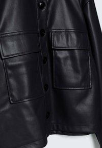 Veste en cuir noir présentant une texture lisse, deux grandes poches à rabat, et six boutons ronds noirs sur le devant.