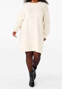 Kreeminsininen neulemekko pitkillä hihoilla, missä on timanttinen tekstuurikuvio bodysuorassa ja rento siluetti. Päällä mustat nilkkurit.