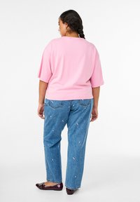 Laisvalaikio rožinės spalvos medvilnės marškinėliai derinami su mėlynomis džinsinėmis kelnėmis, papuoštomis gėlių siuvinėjimu. Subjektas turi nerta šukuosena ir vilki tamsius batus.