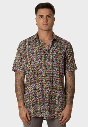 Hombre con cabello corto y oscuro que lleva una camisa de manga corta y colores llamativos con patrones abstractos, de pie frente a un fondo liso.