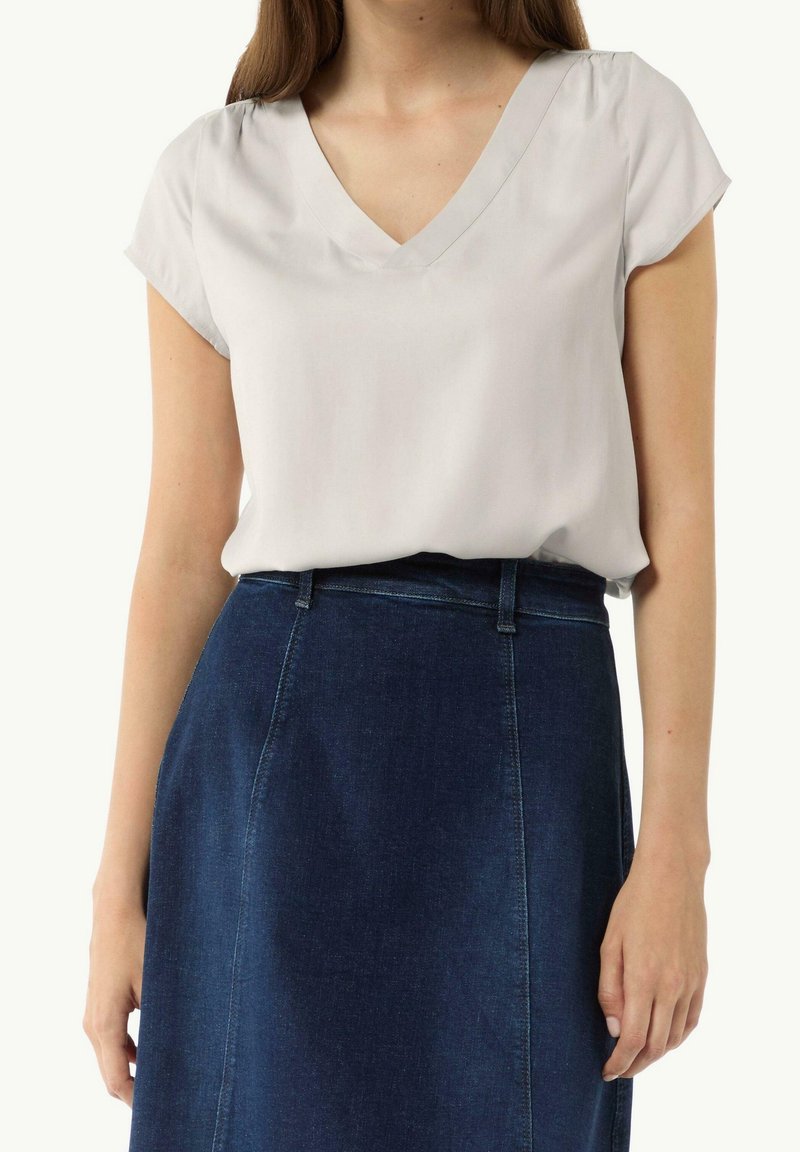 Femme portant un chemisier blanc à manches courtes et col en V, rentré dans une jupe en jean bleu foncé taille haute, debout avec les bras détendus.