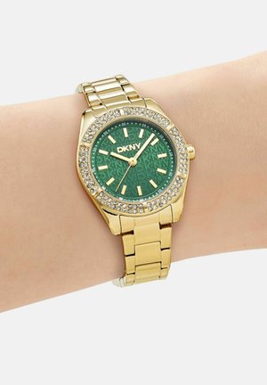 Montre bracelet DKNY ton or avec cadran vert, lunette ornée de cristaux et index dorés sur un bracelet métallique à maillons.
