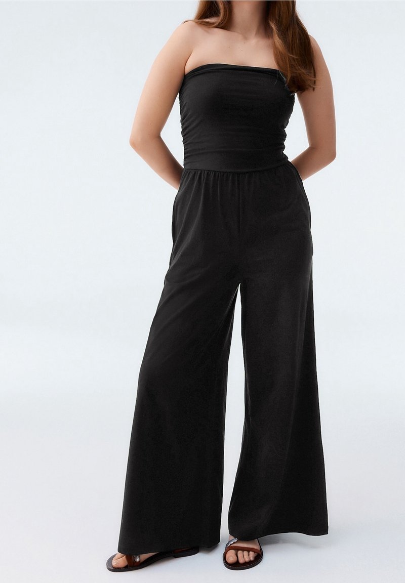 Femme portant une combinaison noire sans bretelles avec un pantalon à jambes larges et des sandales noires, debout les mains derrière le dos sur un fond uni.
