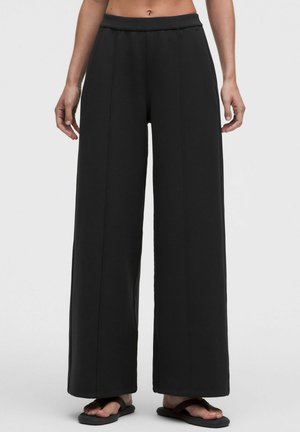 Donna che indossa pantaloni neri a vita alta e gamba larga con cuciture frontali e infradito nere, in piedi davanti a uno sfondo chiaro e uniforme.