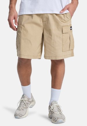 Beige Cargoshorts mit weiter Passform, mehreren Taschen und robustem Baumwollmaterial. Kombiniert mit weißen Socken und hellgrauen Turnschuhen.