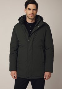 Mężczyzna w ciemnozielonej zimowej kurtce z kapturem zapinanej na zamek, noszący czarną koszulę, stojący na tle jednolitego, neutralnego koloru.