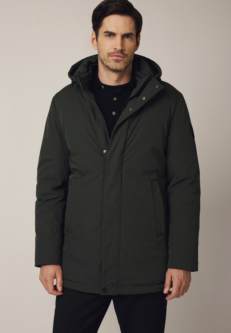 Mężczyzna w ciemnozielonej zimowej kurtce z kapturem zapinanej na zamek, noszący czarną koszulę, stojący na tle jednolitego, neutralnego koloru.