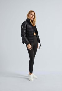 Sort quilted pufferjakke med høj krave, lynlåslukning og logoapplikation. Parret med sorte leggings og hvide sneakers.