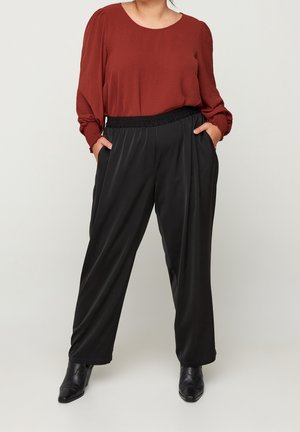 Woman wearing a rust-colored long-sleeve blouse, black high-waisted wide-leg pants, and black ankle boots, hands in pockets, standing.