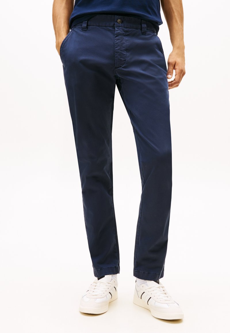 Άνδρας που φοράει σκούρο μπλε στενά παντελόνια και λευκά αθλητικά παπούτσια, στέκεται με τα χέρια στις τσέπες μπροστά σε λευκό φόντο.