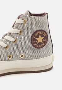 Visoke All Star Converse teniske iz bež platna z maroon podrobnostmi, ki imajo zlate luknje za vezalke, bele vezalke in oznako logotipa.