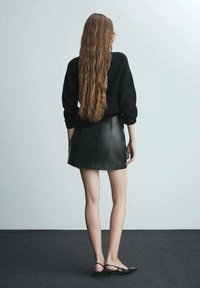 Maglione nero lavorato a maglia, gonna corta in pelle nera, capelli lunghi e ondulati, scarpe nere con tacco sottile, in piedi su un tappeto grigio davanti a uno sfondo chiaro.