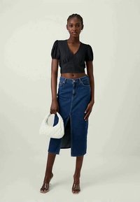 Schwarzes Cropped-Top mit Puffärmeln, blauer Denim-Rock mit Schlitz vorne, durchsichtige Sandalen mit Absatz und weiße gesteppte Handtasche. Neutraler Hintergrund.