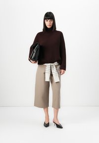 Frau in einem dunkelbraunen Rippstrickpullover, beigen, kurzen, weiten Hosen mit gebundenem gestreiften Hemd in der Taille, schwarzen High Heels und einer schwarzen Clutch.
