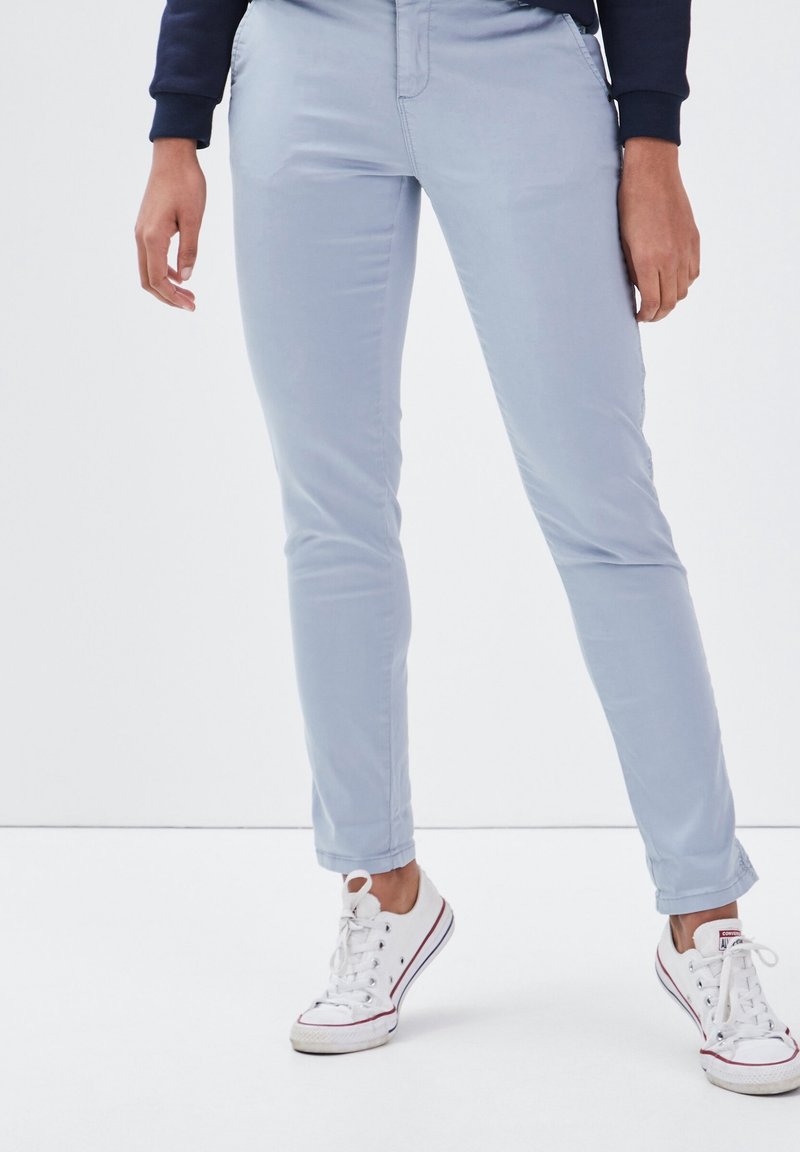 Personne portant un pantalon slim bleu clair et des baskets basses blanches à lacets blancs, debout devant un fond blanc uni.