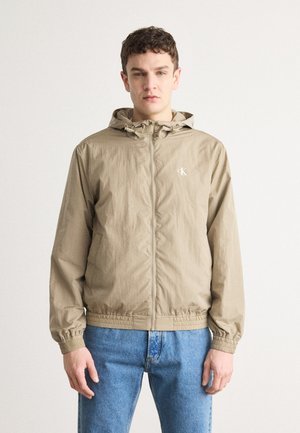Beige zip-up jacket with a hood, elastic cuffs, and drawstring hem. Features a subtle logo on the chest and lightweight, textured fabric.
