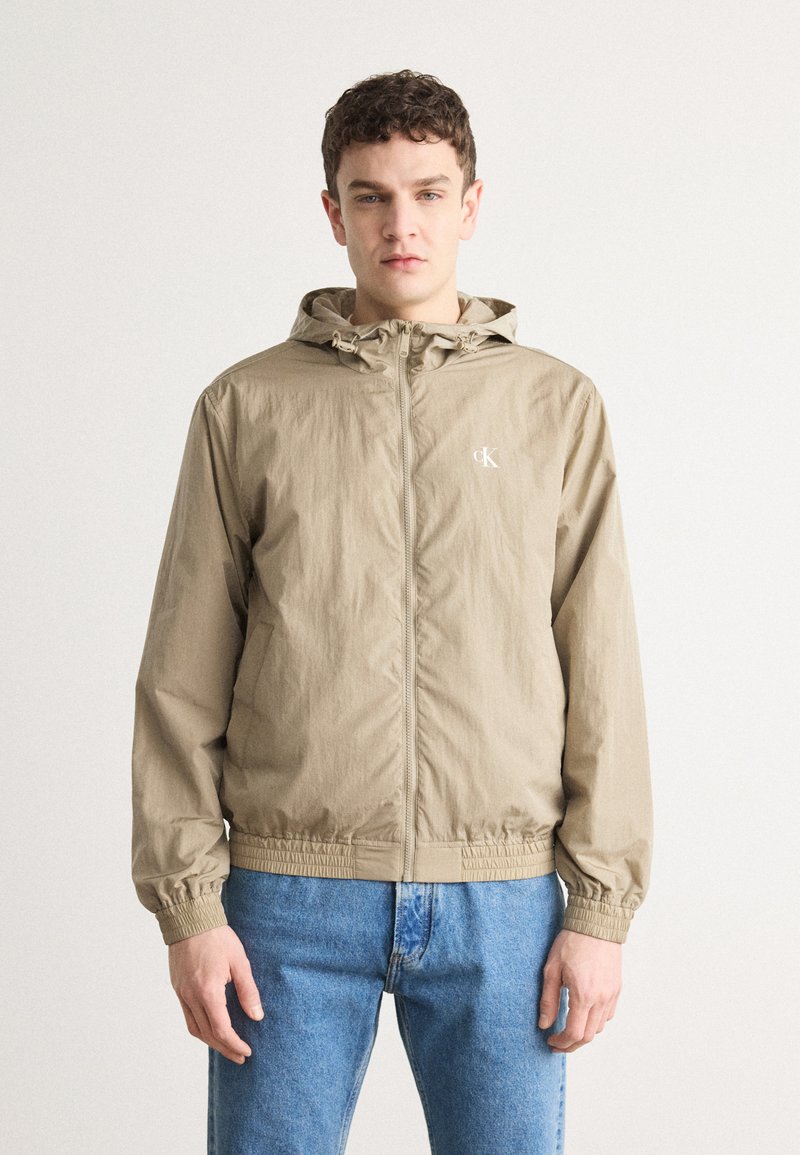 Beige zip-jakke med hætte, elastiske manchetter og snor i bunden. Har et diskret logo på brystet og letvægts, tekstureret stof.