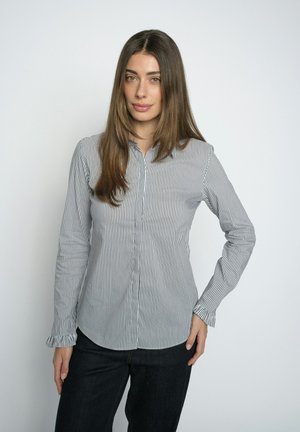 Femme aux longs cheveux bruns portant une chemise blanche ajustée à manches longues avec des rayures verticales noires et un pantalon noir, debout avec la main sur la hanche.