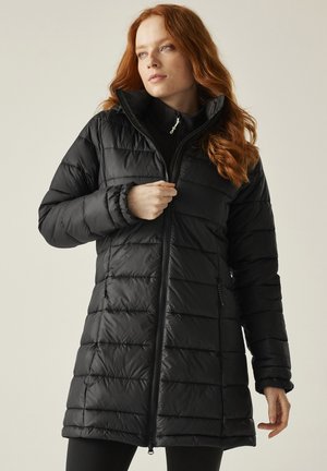 Mujer con cabello rojo cerrándose un abrigo negro acolchado con cuello alto, de pie frente a un fondo claro y liso.