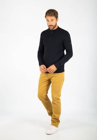 Maglione zip-up blu scuro con collo e polsini a costine, abbinato a chinos beige chiaro e scarpe da ginnastica bianche. Il modello sta in piedi con le mani incrociate.