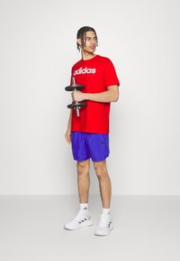 Sarkana kokvilnas T-kreklā ar "adidas" logo, saskaņota ar zilām šortiem. Individuāls tur melnu hanteli, valkājot baltus sporta apavus.