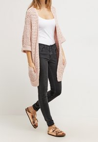 Kobieta w białym topie na ramiączkach, otwartej różowej dzianinowej kardiganie, czarnych dopasowanych dżinsach oraz metalicznych sandałach w odcieniu różowego złota z podwójnymi paskami, idąca.