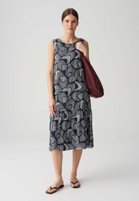 Ärmelloses, marineblaues Kleid mit weißem abstraktem Muster, gestuftem Saum und lockerer Passform. Das Model hält eine bordeauxfarbene Tasche und trägt schwarze Flip-Flops.