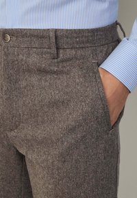 Brązowe spodnie z tkaniny tweed pokryte fakturą, z bocznymi kieszeniami i zapięciem na guzik, w zestawieniu z jasnoniebieską koszulą w paski.
