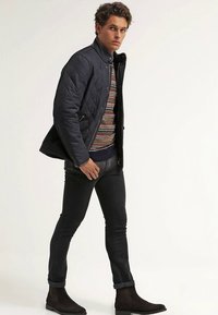 Navy quilted jacket over a patterned sweater, paired with black jeans and dark suede ankle boots. Model poses with one hand in pocket.