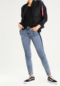 Veste bomber noire avec une tirette de fermeture éclair rouge, associée à un jean skinny bleu clair et des baskets noires à semelles blanches. Tenue simple et décontractée.