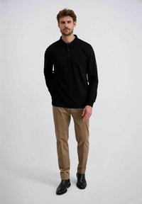 Ein Mann trägt ein schwarzes Langarm-Polo, beige Cordhosen und schwarze Lederstiefel und steht vor einem schlichten hellen Hintergrund.