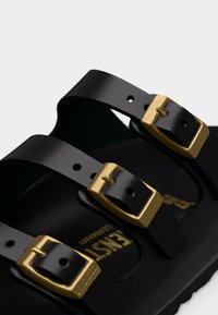 Μαύρο σανδάλι με τρεις ματ λουριά και χρυσά μεταλλικά αγκράφες, που δείχνει μέρος της υφής της σόλας και το ανάγλυφο λογότυπο στο εσωτερικό.