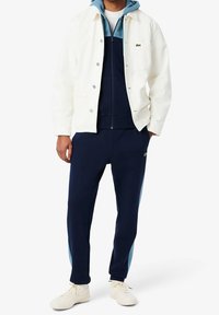 Veste en denim blanc portée sur un survêtement bleu marine comprenant un sweat à capuche zippé bleu. Baskets blanches complètent le look. Logo Lacoste sur la veste et le pantalon.