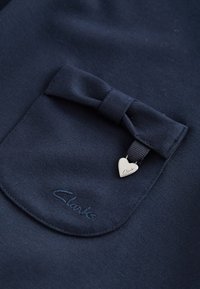 Marineblå stoflomme med en matchende sløjfe og et lille sølv hjerteformet mærke graveret med "Clarks".