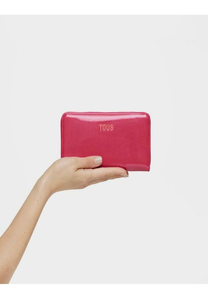 Cartera de cuero brillante rosa sostenida en una mano, con cierre de cremallera y logotipo "TOUS" en relieve en la parte frontal. Textura suave, forma rectangular.