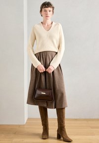 Κρέμα ριμπ πουλόβερ, καφέ πλισέ φούστα, καφέ σουέτ μπότες μέχρι το γόνατο και καφέ τσάντα χειρός με χρυσή λεπτομέρεια.