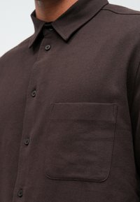 Camicia marrone a maniche lunghe con una texture morbida, colletto strutturato e una tasca sul petto. I bottoni neri creano un contrasto con il tessuto.