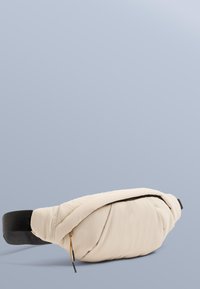 Beige nylonmidjeväska med texturerad yta, böjd design och svart justerbart band. Har ett blixtlåsförsett fack och minimalistiska detaljer.